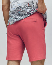 A person wearing Psycho Bunny’s MENS SHIRO KNIT HONEYCOMB SHORT - B6R585F200 in coral pink and a white graphic tee stands with one hand in their pocket, showcasing the back view of the honeycomb-patterned shorts.