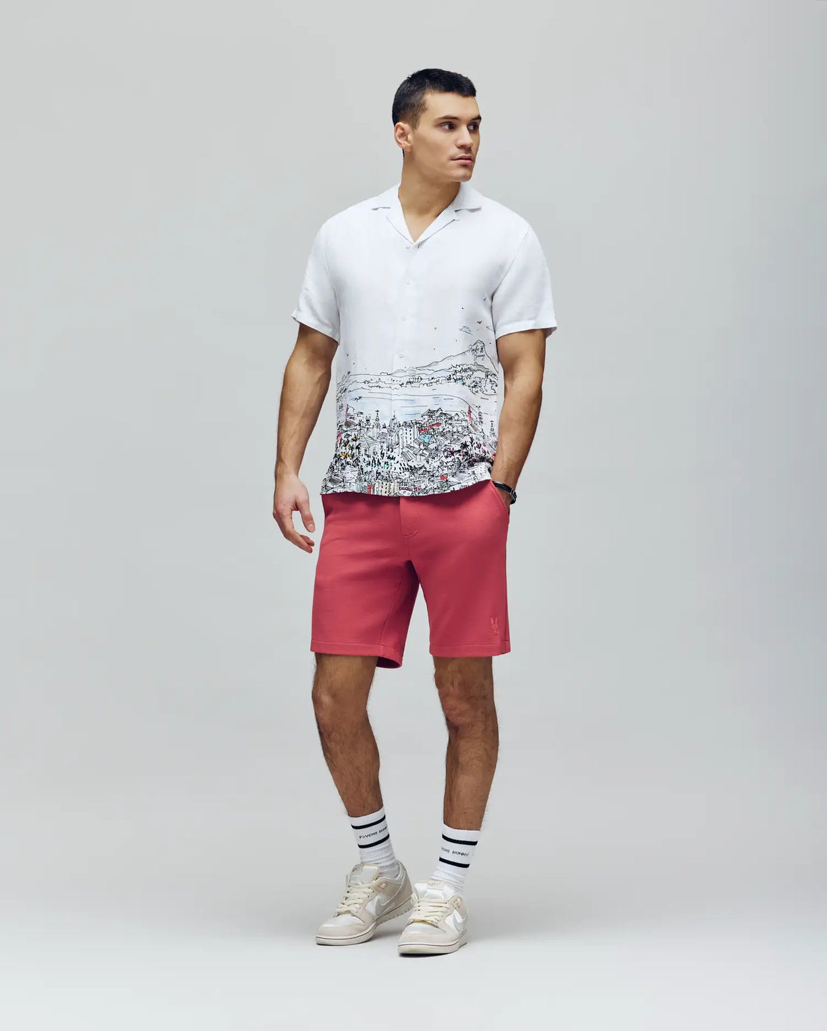 A man stands against a plain background wearing a white cityscape print shirt, coral pink Psycho Bunny MENS SHIRO KNIT HONEYCOMB SHORT - B6R585F200, white sneakers, and white socks with black stripes. He looks to the side neutrally.