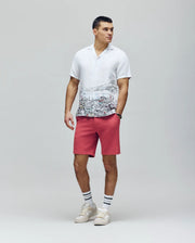 A man stands against a plain background wearing a white cityscape print shirt, coral pink Psycho Bunny MENS SHIRO KNIT HONEYCOMB SHORT - B6R585F200, white sneakers, and white socks with black stripes. He looks to the side neutrally.