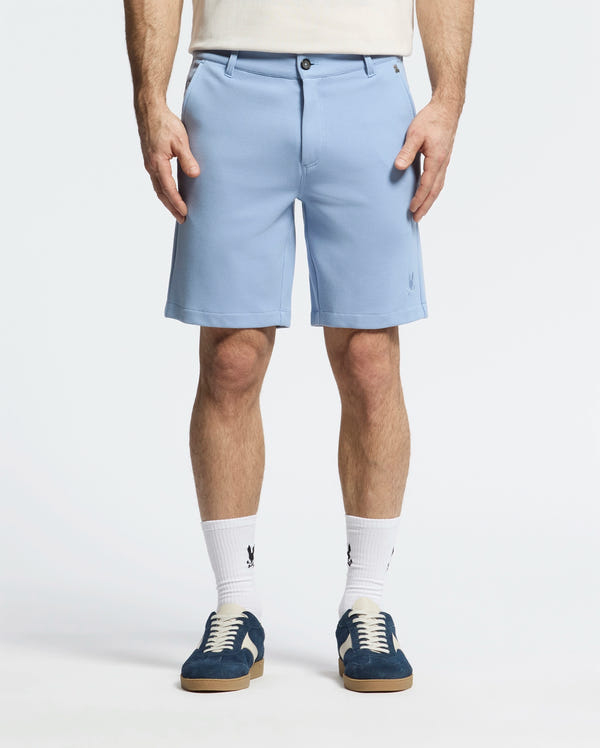 A person is wearing Psycho Bunny's MENS SHIRO KNIT HONEYCOMB SHORT - B6R585F200 in light blue with a honeycomb pattern, white socks, and dark blue sneakers. Standing against a plain background, both hands are in their pockets, showcasing the shorts' embroidered bunny logo on the left.