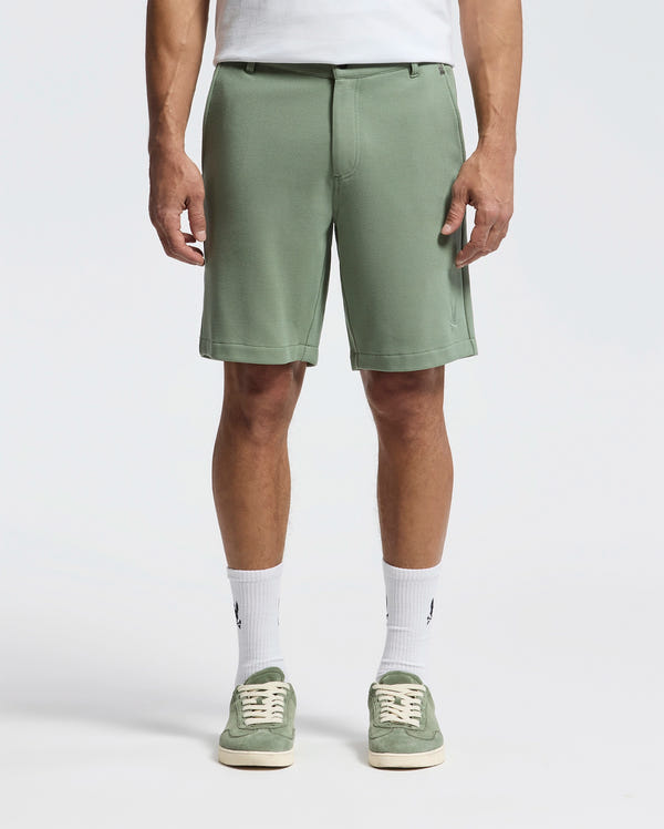 The image shows a person in Psycho Bunny's MENS SHIRO KNIT HONEYCOMB SHORT (B6R585F200) in sage green with a white T-shirt tucked in, honeycomb-patterned piqué crew socks, and matching green sneakers against a plain white backdrop—perfect for any casual outing.