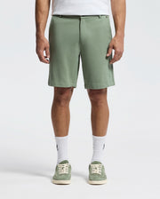 The image shows a person in Psycho Bunny's MENS SHIRO KNIT HONEYCOMB SHORT (B6R585F200) in sage green with a white T-shirt tucked in, honeycomb-patterned piqué crew socks, and matching green sneakers against a plain white backdrop—perfect for any casual outing.