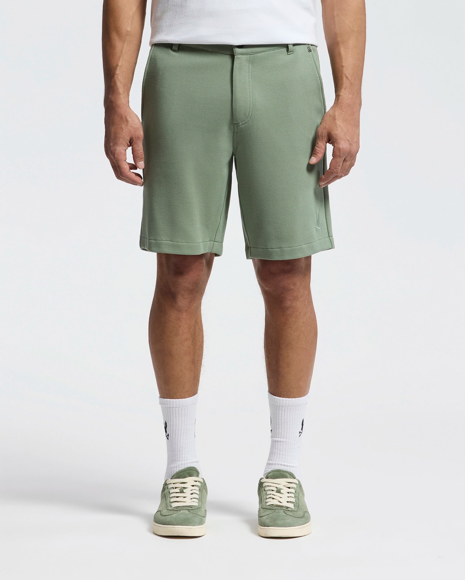 The image shows a person in Psycho Bunny's MENS SHIRO KNIT HONEYCOMB SHORT (B6R585F200) in sage green with a white T-shirt tucked in, honeycomb-patterned piqué crew socks, and matching green sneakers against a plain white backdrop—perfect for any casual outing.