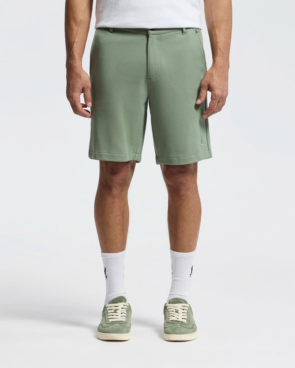 The image shows a person in Psycho Bunny's MENS SHIRO KNIT HONEYCOMB SHORT (B6R585F200) in sage green with a white T-shirt tucked in, honeycomb-patterned piqué crew socks, and matching green sneakers against a plain white backdrop—perfect for any casual outing.