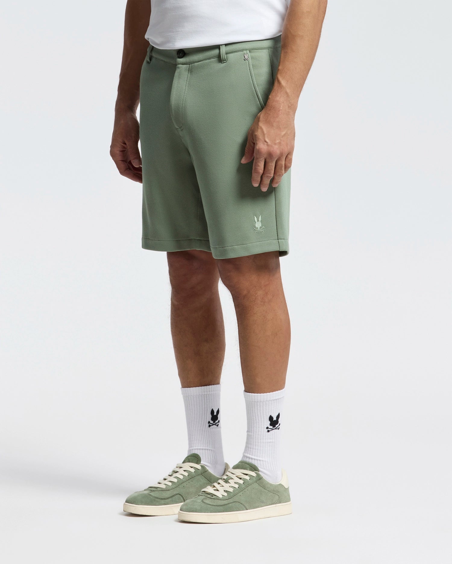 A person is wearing Psycho Bunny's sage green MENS SHIRO KNIT HONEYCOMB SHORTS paired with white socks featuring a black logo and green sneakers with white soles. The honeycomb-patterned piqué fabric adds texture, all set against a plain white background.
