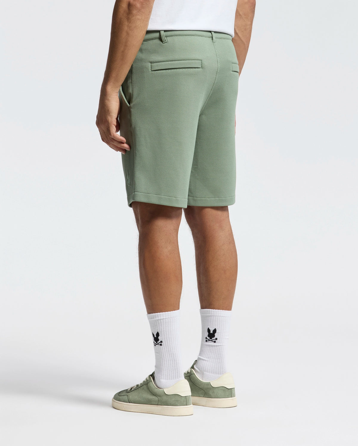 The person is wearing versatile sage green MENS SHIRO KNIT HONEYCOMB SHORT - B6R585F200 by Psycho Bunny, paired with a white shirt and socks featuring the black bunny logo. Sage green sneakers and hands tucked in pockets complete their casually cool look as they face away.