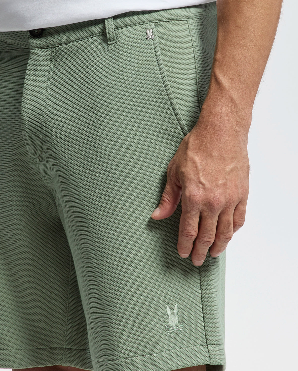 A person is wearing Psycho Bunny's MENS SHIRO KNIT HONEYCOMB SHORT, B6R585F200. These green shorts feature a honeycomb pattern and a rabbit logo on the hem. The background is plain white, and their right hand rests relaxed at their side.