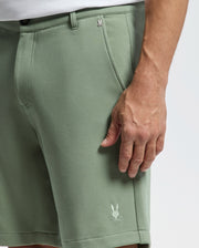 A person is wearing Psycho Bunny's MENS SHIRO KNIT HONEYCOMB SHORT, B6R585F200. These green shorts feature a honeycomb pattern and a rabbit logo on the hem. The background is plain white, and their right hand rests relaxed at their side.