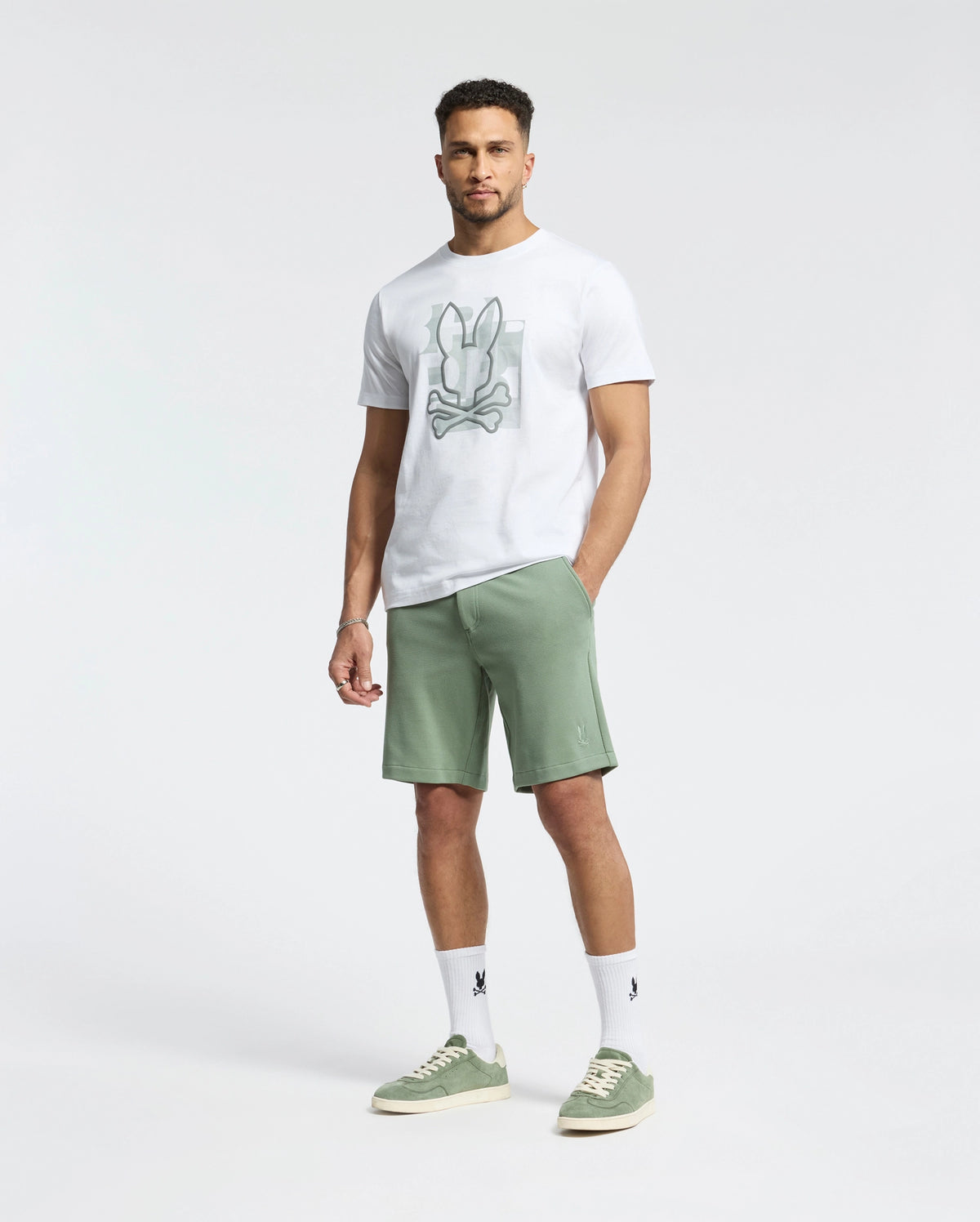 A man stands against a white background in a honeycomb-patterned piqué T-shirt paired with Psycho Bunny's MENS SHIRO KNIT HONEYCOMB SHORT - B6R585F200 in light green, green sneakers, and white socks. His short curly hair complements his neutral expression, offering versatility for any wardrobe.