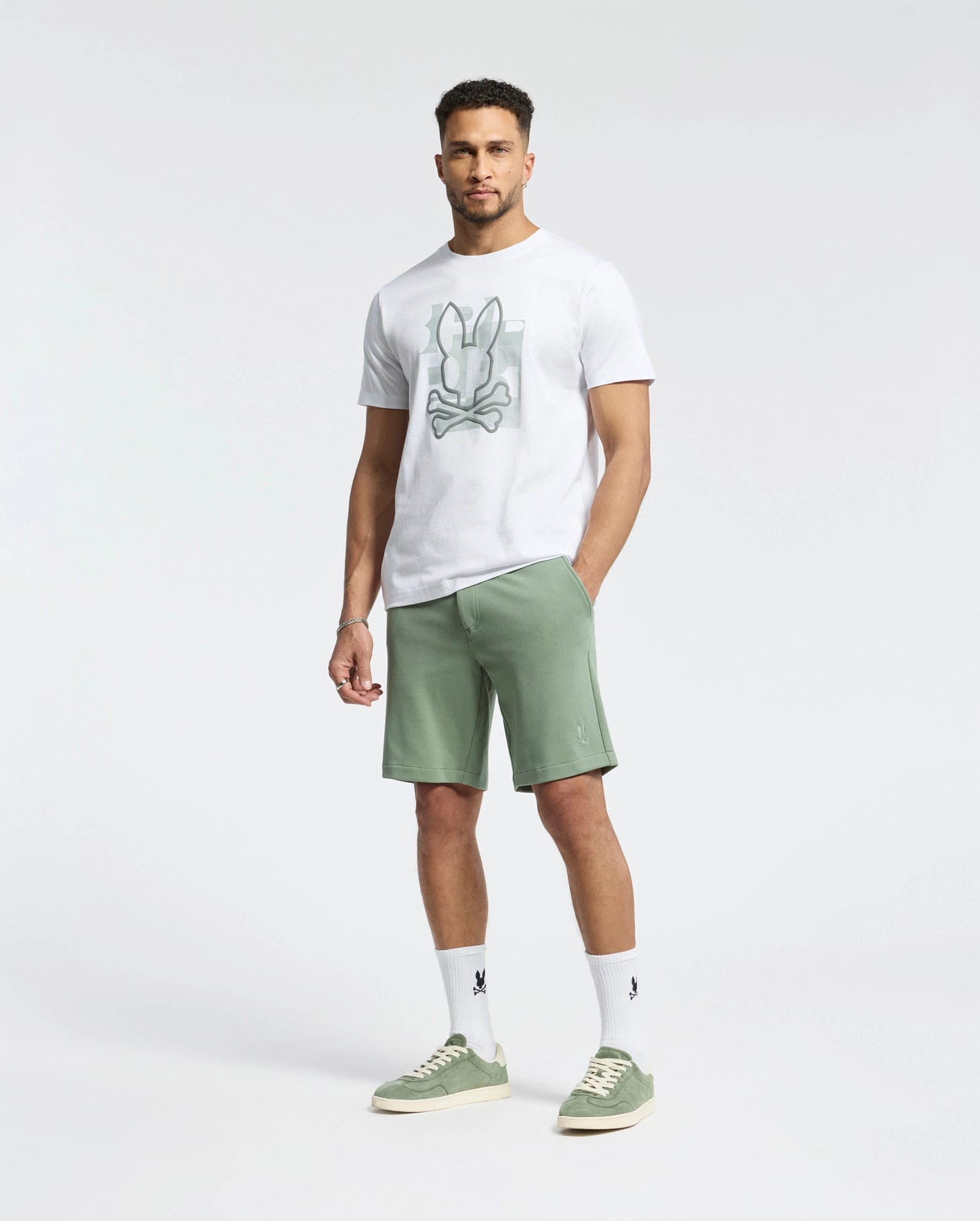 A man stands against a white background in a honeycomb-patterned piqué T-shirt paired with Psycho Bunny's MENS SHIRO KNIT HONEYCOMB SHORT - B6R585F200 in light green, green sneakers, and white socks. His short curly hair complements his neutral expression, offering versatility for any wardrobe.