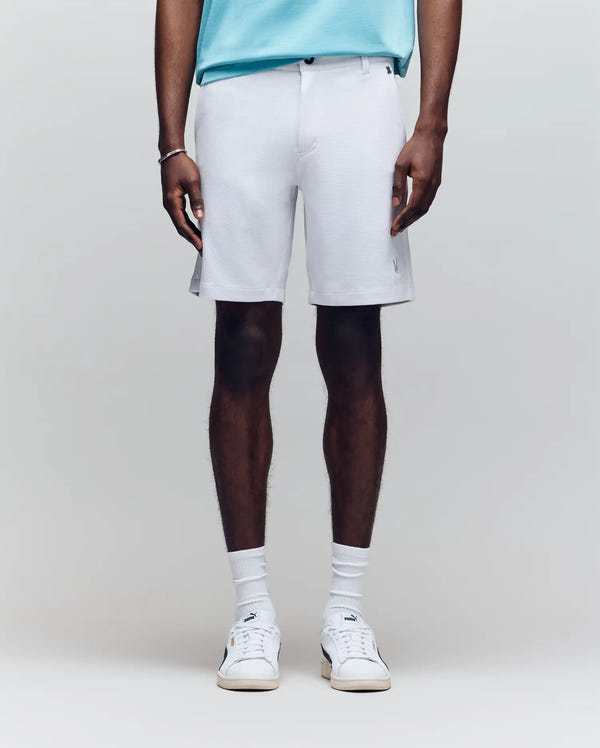 A person stands against a plain light background, shown from shoulders to feet, wearing a light blue honeycomb-patterned piqué shirt and Psycho Bunny MENS SHIRO KNIT HONEYCOMB SHORT - B6R585F200 in white, with white crew socks and sneakers.