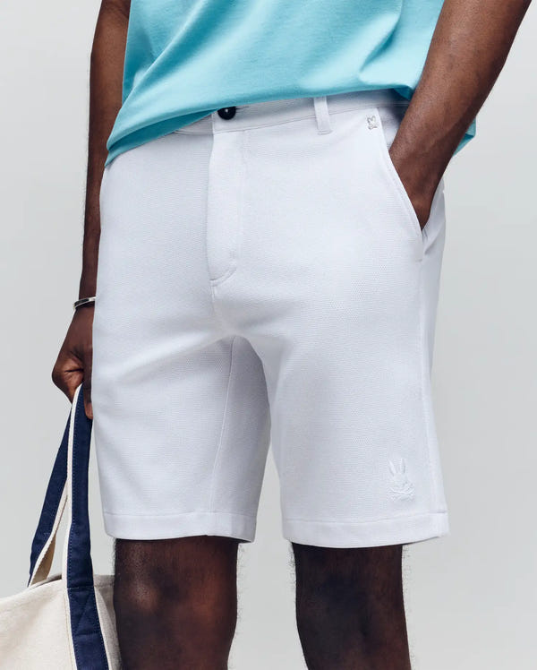 Wearing Psycho Bunny's light gray MENS SHIRO KNIT HONEYCOMB SHORTS, a person pairs them with a light blue honeycomb piqué shirt and holds a beige and navy tote, standing against a neutral background, one hand in their pocket.