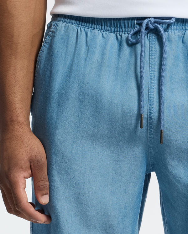 A person is shown wearing Psycho Bunny's MENS KERVIN LIGHTWEIGHT DENIM WOVEN SHORT (B6R633F200) in light blue, featuring an elasticized waist and drawstring. They complement the shorts with a white top, casually resting their left hand by their side.