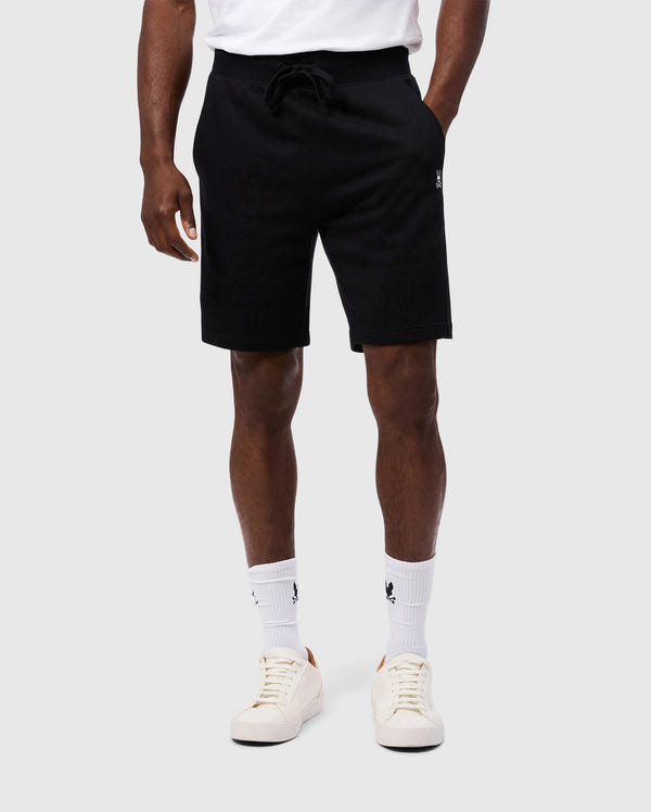A person wearing a white t-shirt, the Psycho Bunny MENS LEVY FLEECE SWEATSHORT (B6R862D200) in black, white socks, and white sneakers stands against a plain grey background. The person's hands are relaxed by their sides, showcasing this everyday staple outfit.