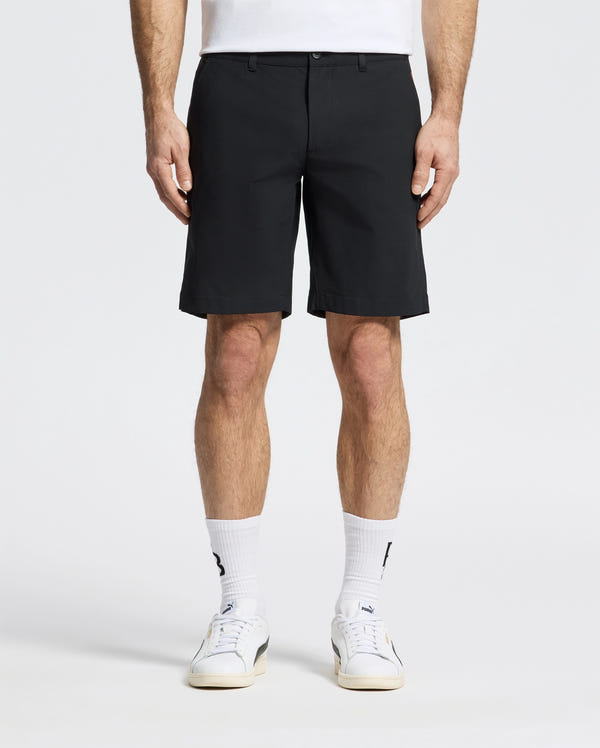A person in Psycho Bunny MENS 10K STEP SHORT - B6R969F200, a white shirt, crew socks, and sneakers stands against a plain light background. The waist-down crop highlights the athletic breathability of the black shorts.