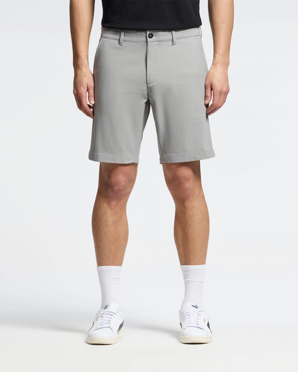 A person wearing Psycho Bunny's MENS 10K STEP SHORT (B6R969F200) stands facing forward in light gray, breathable shorts that reach just above the knees, paired with a black shirt, white sneakers, and white socks against a plain white background.