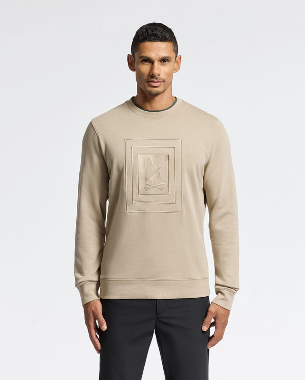 A person poses wearing the Psycho Bunny MENS OAK CITY CREW NECK - B6S274E200, a premium quality beige sweatshirt featuring raised embroidery in an embossed rectangular design on the front, paired with black pants. The men's crew neck sweater is set against a plain white background.