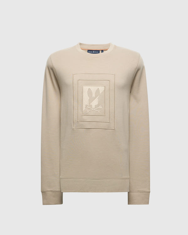 The Psycho Bunny Men's Oak City Crew Neck - B6S274E200 is a premium quality beige long-sleeve sweatshirt featuring raised embroidery of a figure with rabbit ears on the center front. This men's sweater is designed with ribbed cuffs and set against a plain white backdrop.