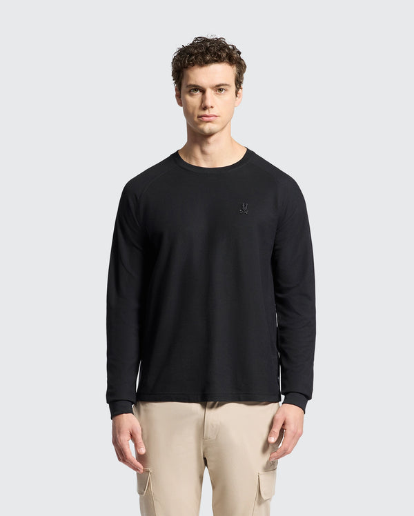 A man with curly hair wears Psycho Bunny's black MENS TAYLOR PIQUE LONG SLEEVE TEE and beige pants, standing against a light gray background. The shirt includes an embroidered Bunny logo on the left chest.
