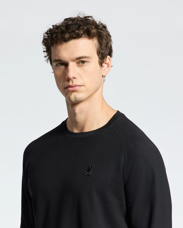 A person with short, curly hair is wearing the Psycho Bunny MENS TAYLOR PIQUE LONG SLEEVE TEE in black Pima cotton piqué. It features a small embroidered bird logo on the chest. The plain white background highlights their stylish look.