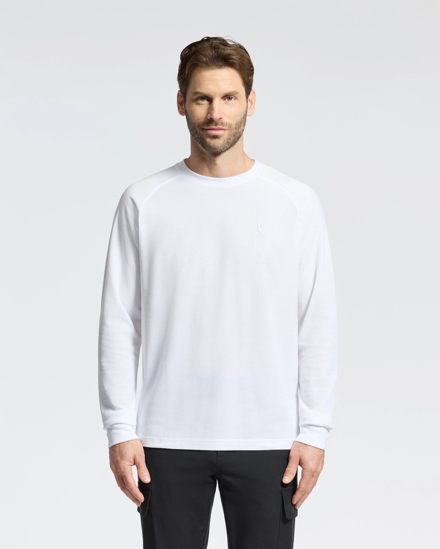 A man with short hair and a beard stands against a plain background, wearing the Psycho Bunny MENS TAYLOR PIQUE LONG SLEEVE TEE in white and black pants. He has a neutral expression, his arms relaxed at his sides, while subtle embroidered bunnies enhance his look.