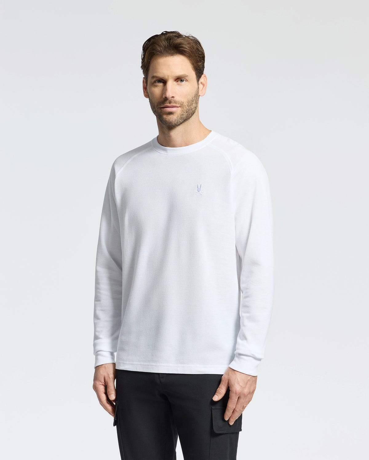 A man with short hair and facial hair wears the Psycho Bunny MENS TAYLOR PIQUE LONG SLEEVE TEE - B6T580F200 in white with raglan sleeves, paired with black pants. He stands against a plain light gray background, slightly facing left, showcasing the embroidered Bunny logo on his chest.