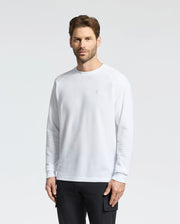A man with short hair and facial hair wears the Psycho Bunny MENS TAYLOR PIQUE LONG SLEEVE TEE - B6T580F200 in white with raglan sleeves, paired with black pants. He stands against a plain light gray background, slightly facing left, showcasing the embroidered Bunny logo on his chest.