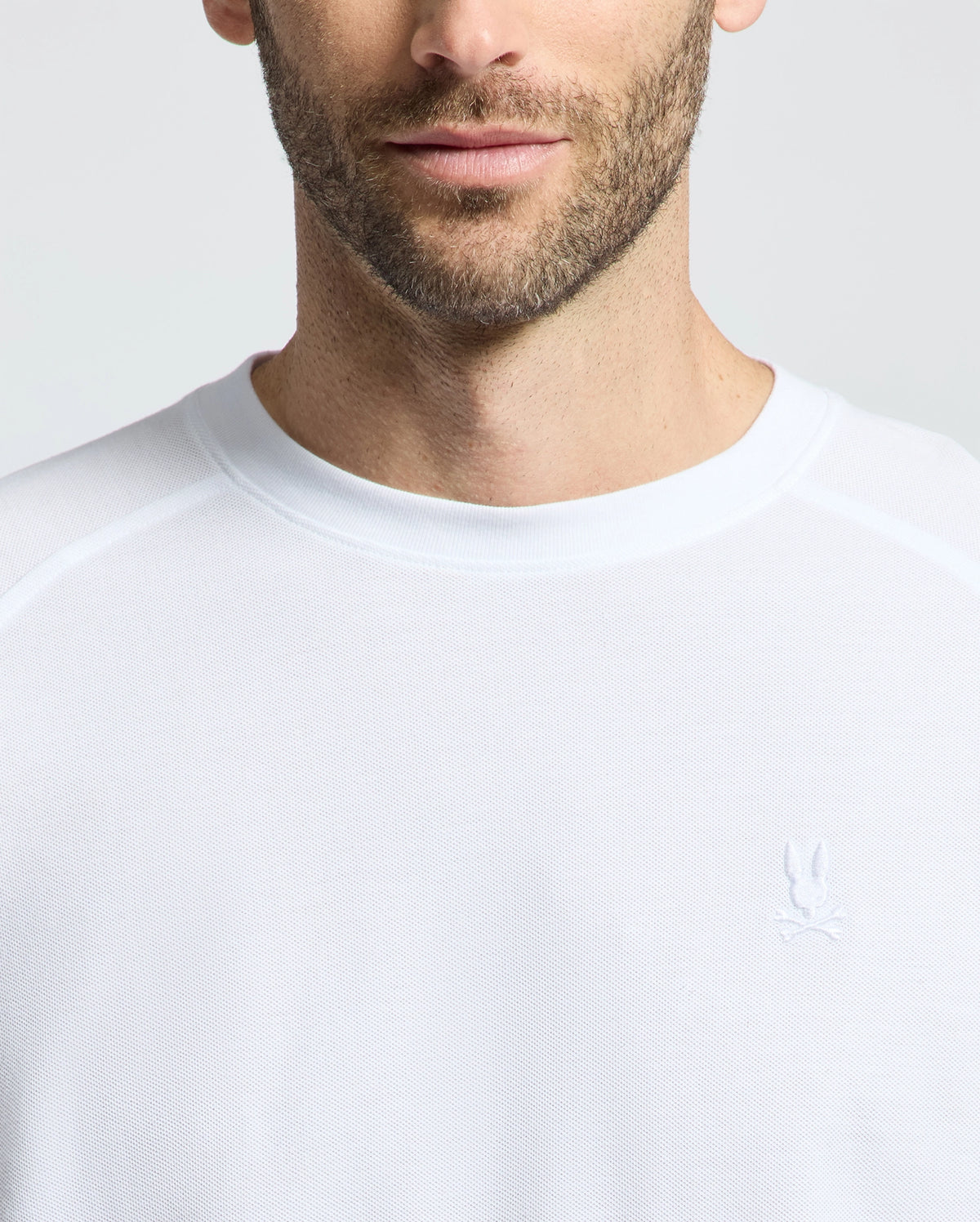 A man wears the Psycho Bunny MENS TAYLOR PIQUE LONG SLEEVE TEE (B6T580F200), a white crew neck shirt made from luxurious Pima cotton, with a small embroidered Bunny logo on the chest, set against a plain light grey background.