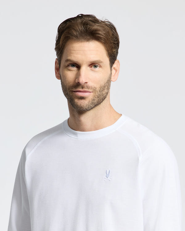 A man with short brown hair and a beard wears the Psycho Bunny Men's Taylor Pique Long Sleeve Tee (B6T580F200), with raglan sleeves and a small embroidered Bunny logo, standing against a plain light background.
