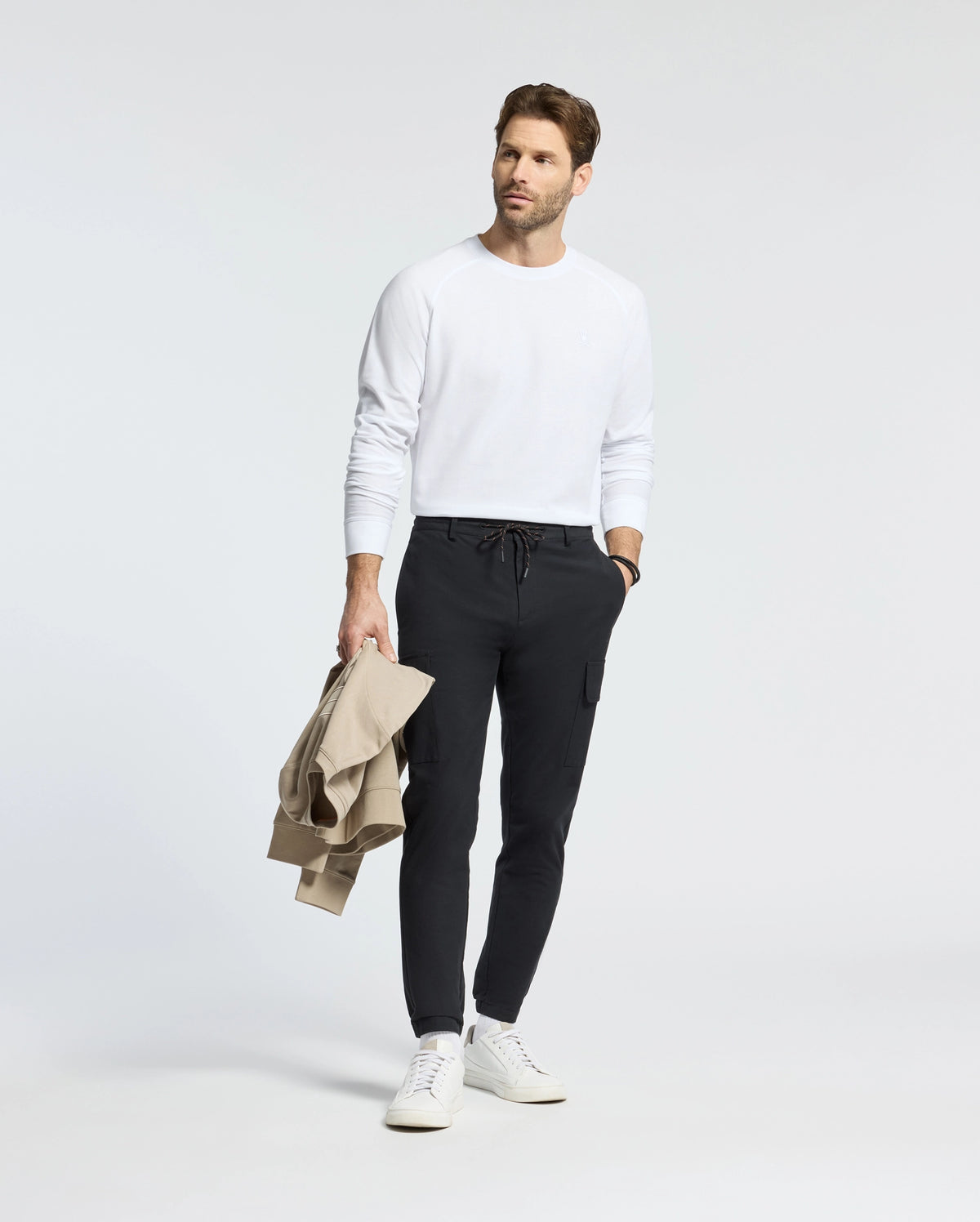 A person wearing a Psycho Bunny MENS TAYLOR PIQUE LONG SLEEVE TEE, dark pants, and white sneakers stands against a plain background holding a beige jacket and sporting a watch, highlighted by the subtle charm of an embroidered Bunny.