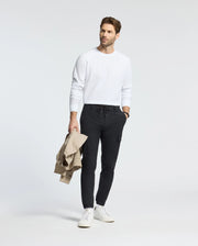 A person wearing a Psycho Bunny MENS TAYLOR PIQUE LONG SLEEVE TEE, dark pants, and white sneakers stands against a plain background holding a beige jacket and sporting a watch, highlighted by the subtle charm of an embroidered Bunny.