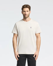 A man with short hair and a beard wears the Psycho Bunny MENS CLASSIC CREW NECK TEE - B6U014B200 in premium Pima cotton, featuring a logo on the left chest. The plain white tee pairs with black pants as he stands against a light background.