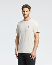 A person with short brown hair and a beard stands against a light gray background, wearing the Psycho Bunny Men's Classic Crew Neck Tee in cream, crafted from premium Pima cotton with an embroidered chest design. Paired with dark pants, they look directly at the camera.