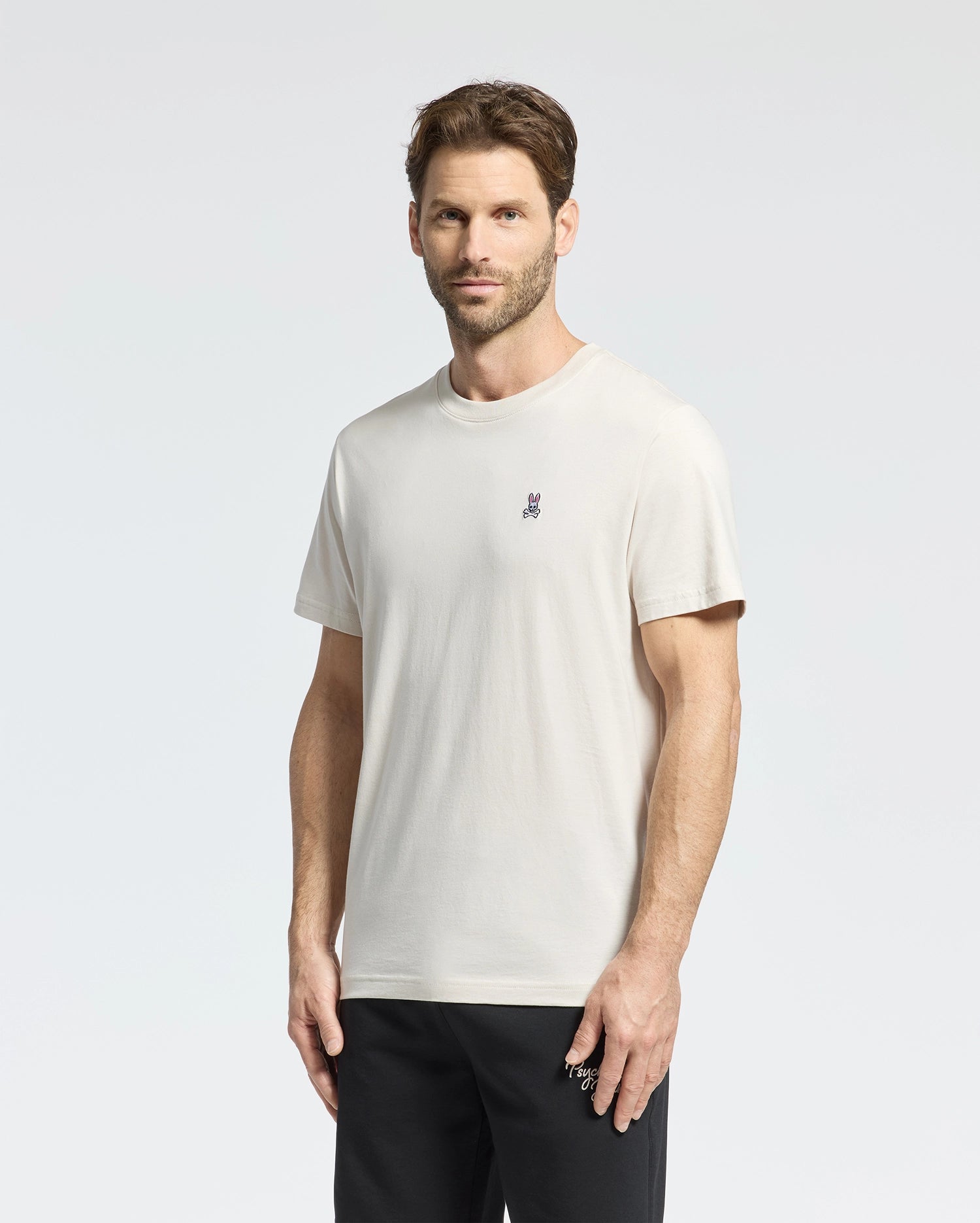 A person with short brown hair and a beard stands against a light gray background, wearing the Psycho Bunny Men's Classic Crew Neck Tee in cream, crafted from premium Pima cotton with an embroidered chest design. Paired with dark pants, they look directly at the camera.