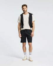 A man stands wearing a Psycho Bunny MENS CLASSIC CREW NECK TEE in white, paired with black shorts and a black jacket tied around his shoulders. He completes the look with striped socks and white sneakers against a plain white background.