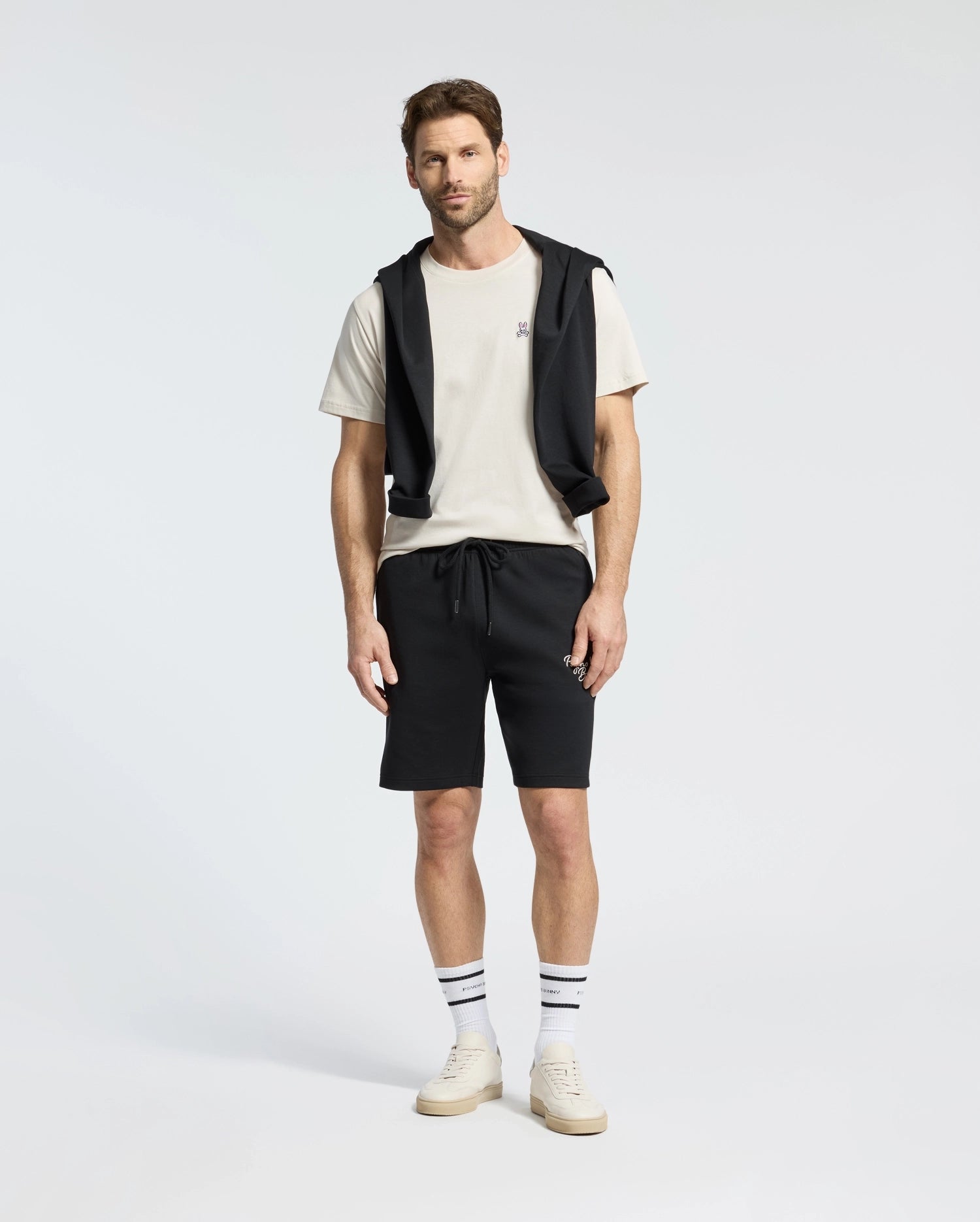 A man stands wearing a Psycho Bunny MENS CLASSIC CREW NECK TEE in white, paired with black shorts and a black jacket tied around his shoulders. He completes the look with striped socks and white sneakers against a plain white background.