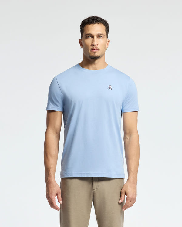 A man with short curly hair and a neutral expression stands against a plain background, wearing Psycho Bunny's MENS CLASSIC CREW NECK TEE (B6U014B200) in light blue with a small embroidered logo on the chest, paired with beige pants.