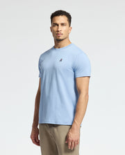 A person in a Psycho Bunny MENS CLASSIC CREW NECK TEE - B6U014B200, light blue with a small embroidered logo, and beige pants stands against a plain white background.