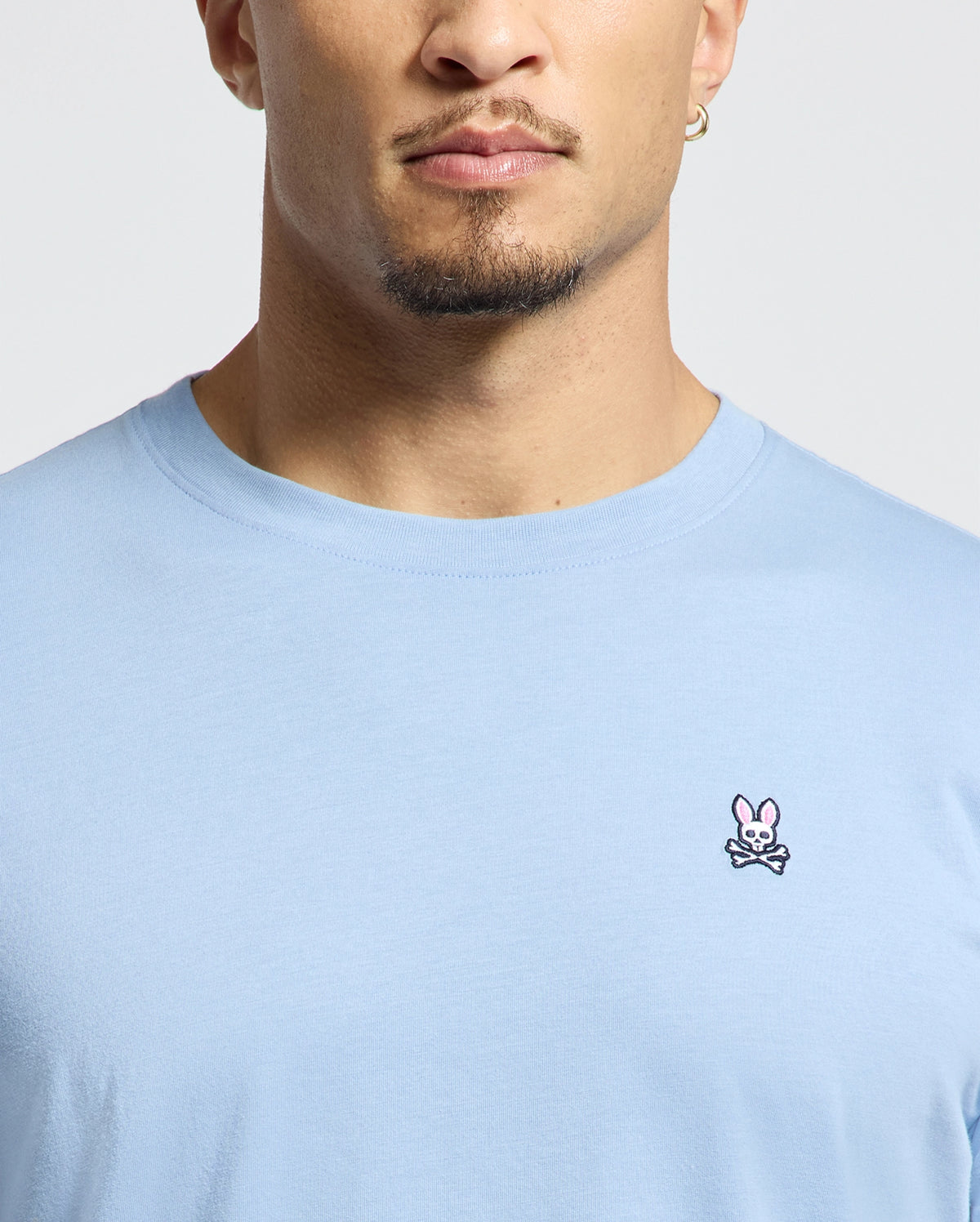 A person is wearing the Psycho Bunny MENS CLASSIC CREW NECK TEE - B6U014B200, a light blue Pima cotton shirt with a pink bunny and crossed bones emblem on the chest. The photo focuses on the lower face and beard against a plain white background, highlighting the tee's subtle yet vibrant design.