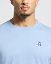 A person is wearing the Psycho Bunny MENS CLASSIC CREW NECK TEE - B6U014B200, a light blue Pima cotton shirt with a pink bunny and crossed bones emblem on the chest. The photo focuses on the lower face and beard against a plain white background, highlighting the tee's subtle yet vibrant design.