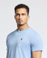 A man with short curly hair and a goatee wears a Psycho Bunny light blue MENS CLASSIC CREW NECK TEE - B6U014B200, crafted from soft Pima cotton with a small embroidered logo on the chest. He has an earring in his left ear and poses against a plain background.