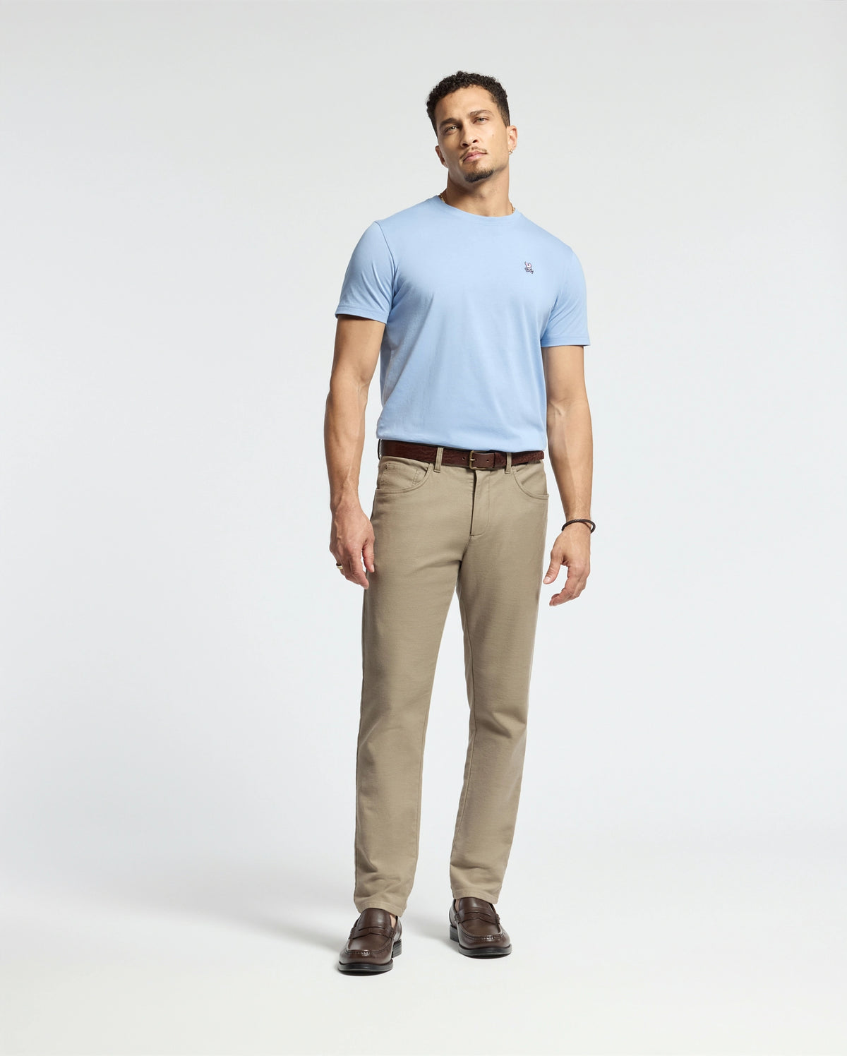 A man stands against a plain background, wearing a light blue Psycho Bunny MENS CLASSIC CREW NECK TEE (B6U014B200), beige pants, and brown loafers. He has short hair and relaxed hands by his sides. The outfit is casual, coordinated, and subtly embraces vibrant colors.