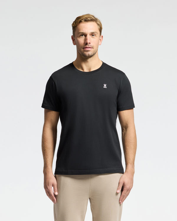 A man is standing against a plain white background, wearing the Psycho Bunny MENS CLASSIC CREW NECK TEE - B6U014CRPC, crafted from 100% Pima cotton with a small embroidered logo on the left chest.