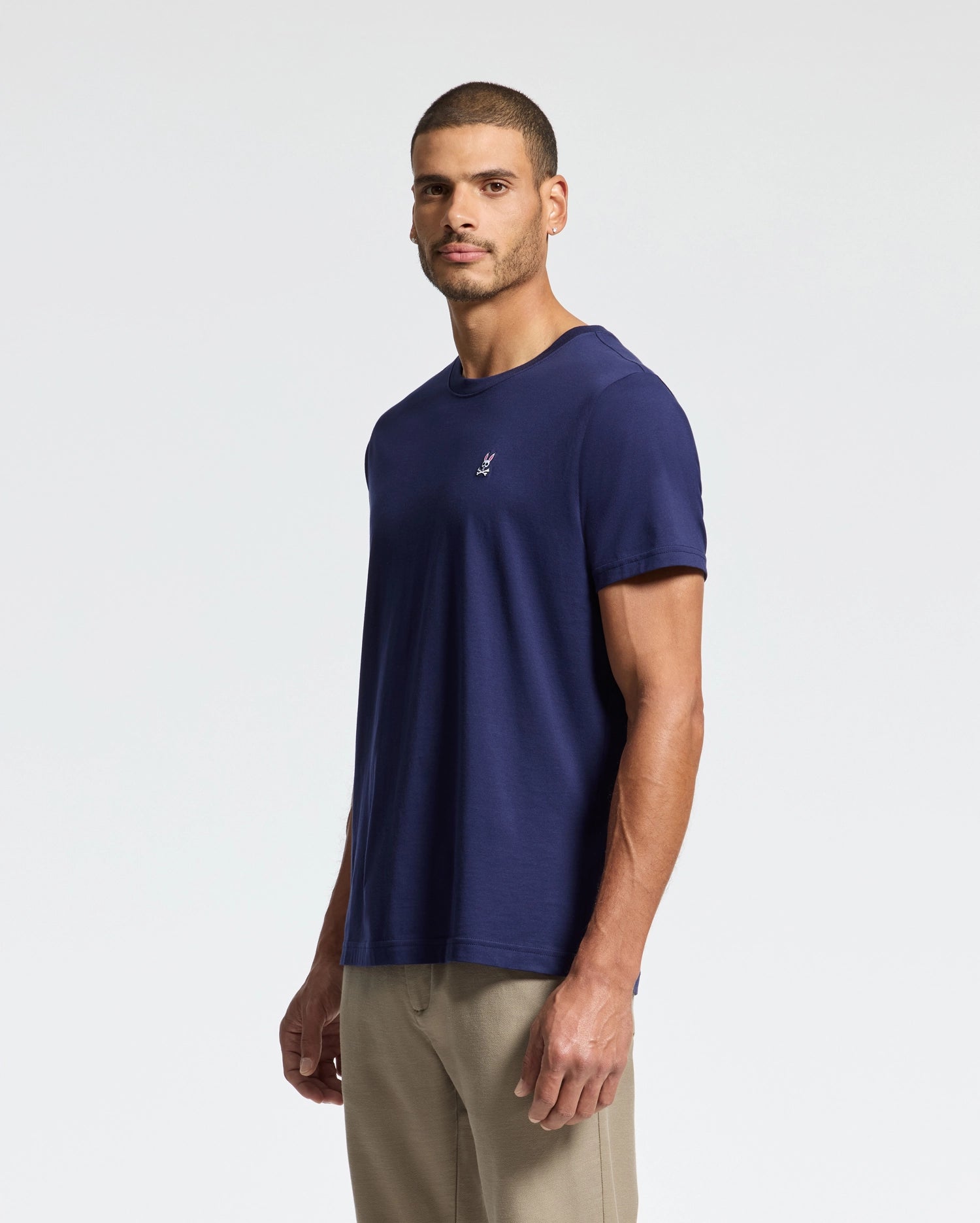 A man stands against a plain background wearing the Psycho Bunny MENS CLASSIC CREW NECK TEE - B6U014CRPC in dark blue and beige pants. This versatile pima cotton t-shirt features a small embroidered logo on the left side. With a neutral expression, he faces the camera, highlighting timeless style and comfort.