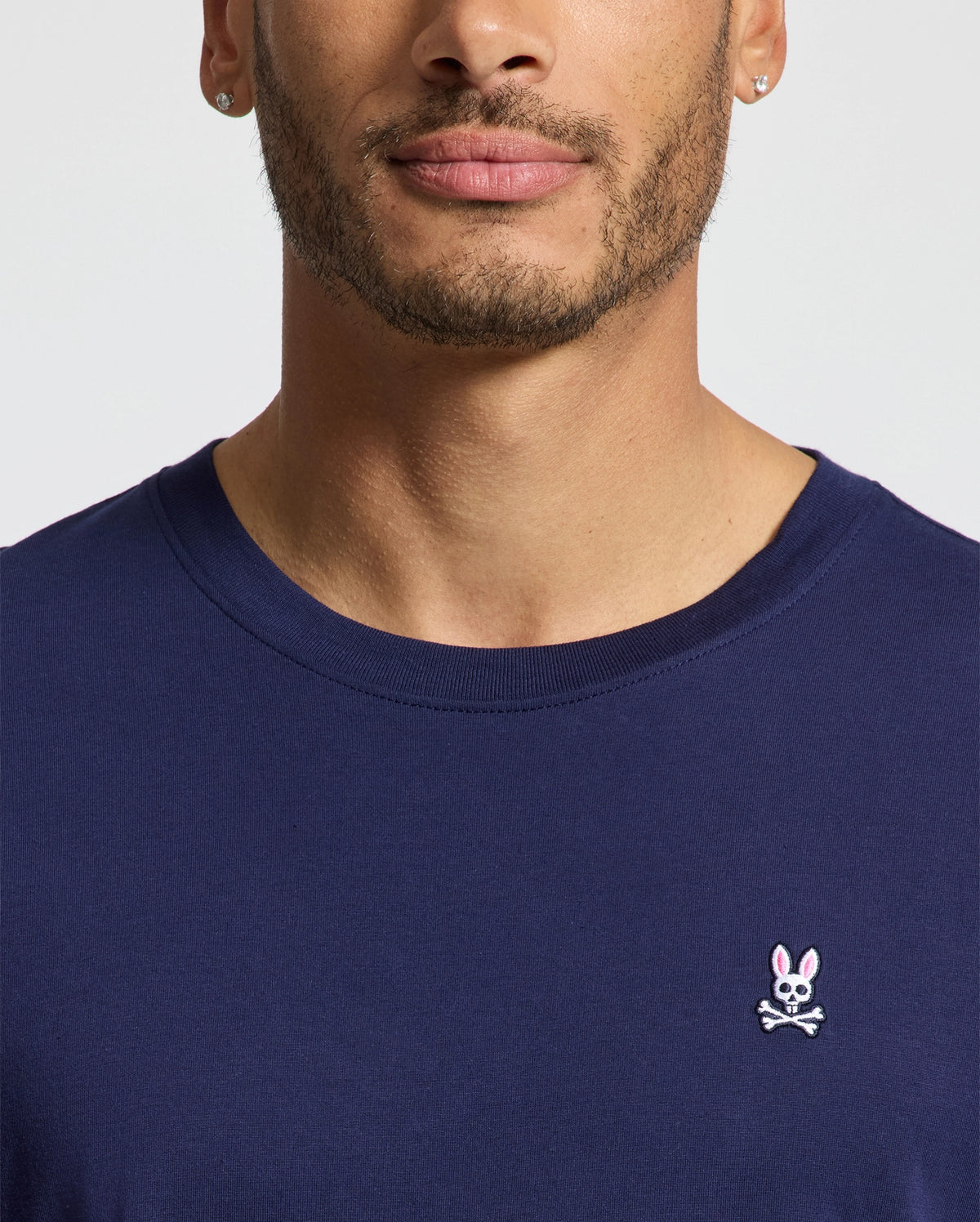 A man wearing the Psycho Bunny MENS CLASSIC CREW NECK TEE - B6U014CRPC in a versatile navy blue, made from soft pima cotton and adorned with a small bunny and crossbones emblem on the left side. The image shows him from the shoulders to just above the mouth, highlighting his short facial hair and small hoop earrings.