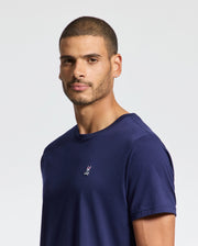 A man with short hair and a beard wears the Psycho Bunny MENS CLASSIC CREW NECK TEE - B6U014CRPC in navy blue pima cotton, which features a small embroidered design on the chest. He gazes slightly to the side with a neutral expression against a plain background, highlighting its versatile style.