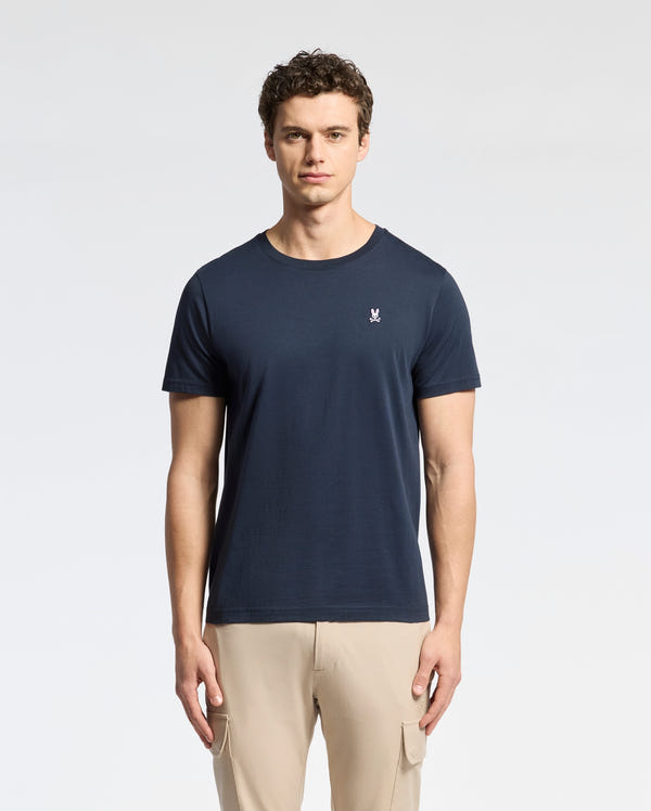 A man wears the Psycho Bunny MENS CLASSIC CREW NECK TEE - B6U014CRPC in navy blue, featuring a small embroidered logo on the left chest and taped seams for comfort. Made from 100% Pima cotton, it pairs perfectly with light beige pants against a plain white background.