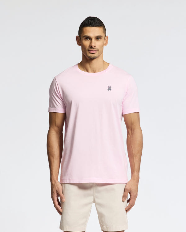 A person with short hair wears a versatile, light pink Psycho Bunny MENS CLASSIC CREW NECK TEE made of soft pima cotton with a small logo on the chest, paired with beige shorts. They stand against a plain white background.
