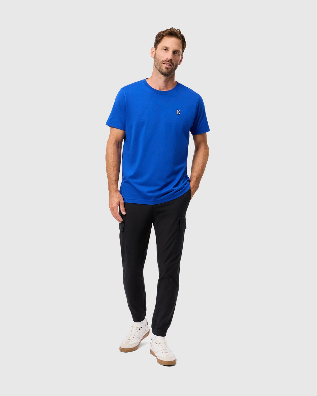 A man in a Psycho Bunny CLASSIC CREW NECK TEE - B6U014CRPC in blue and black jogger pants stands against a plain background. He is wearing white sneakers with a subtle embroidered logo, one hand tucked in his pocket while the other hangs by his side.