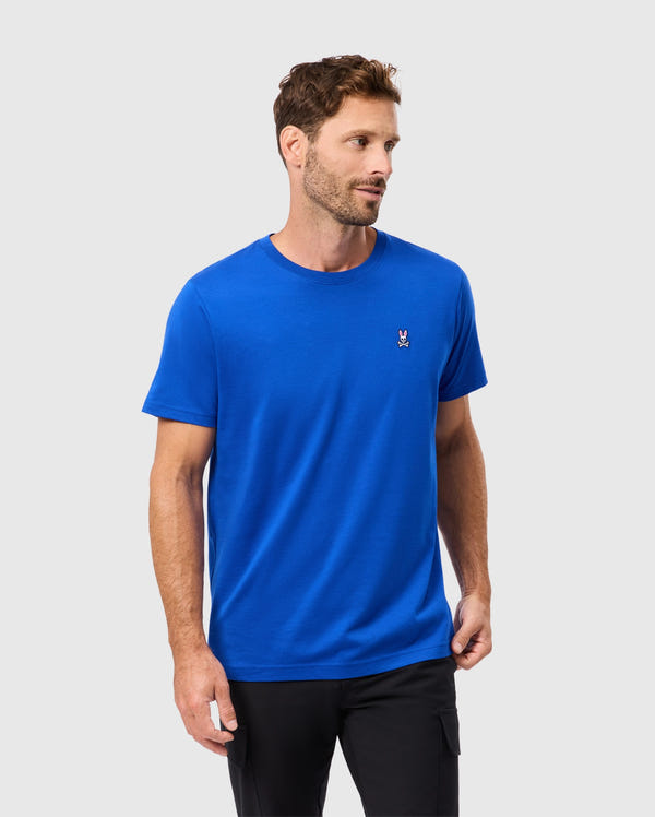 A man with short hair and a beard is wearing a Psycho Bunny MENS CLASSIC CREW NECK TEE in bright blue, crafted from soft Pima cotton, featuring an embroidered logo on the chest. He is looking to the side while wearing black pants against a light gray background.